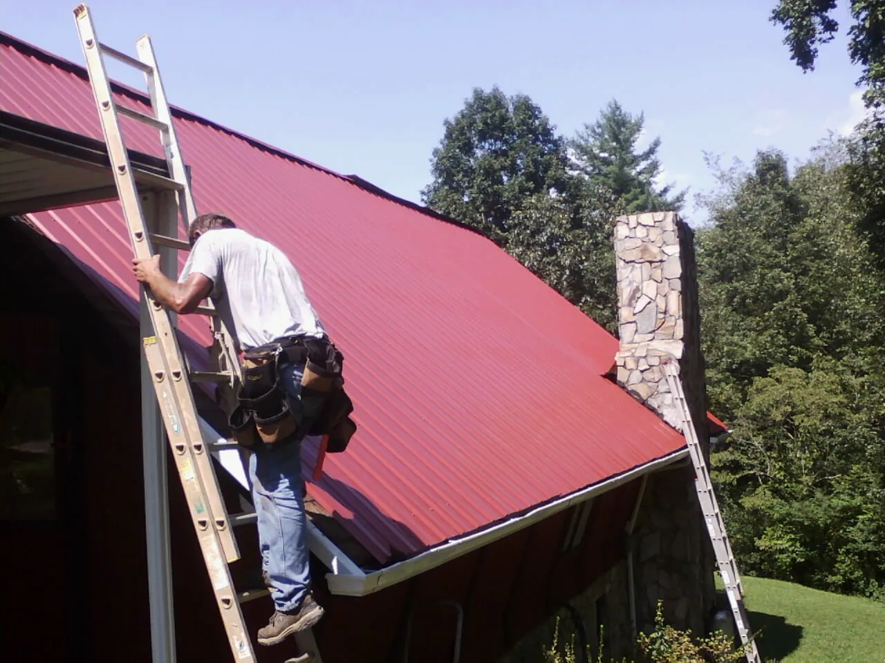 Roofer on a ladder beside a red metal roof and stone chimney — Soffit & Fascia Repair by Roofing Brothers in Wentzville